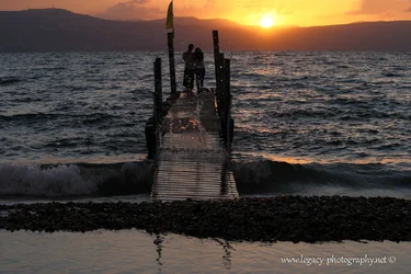 $Couple standing on a dock on the Kinneret at sunset with wave splash.webp
